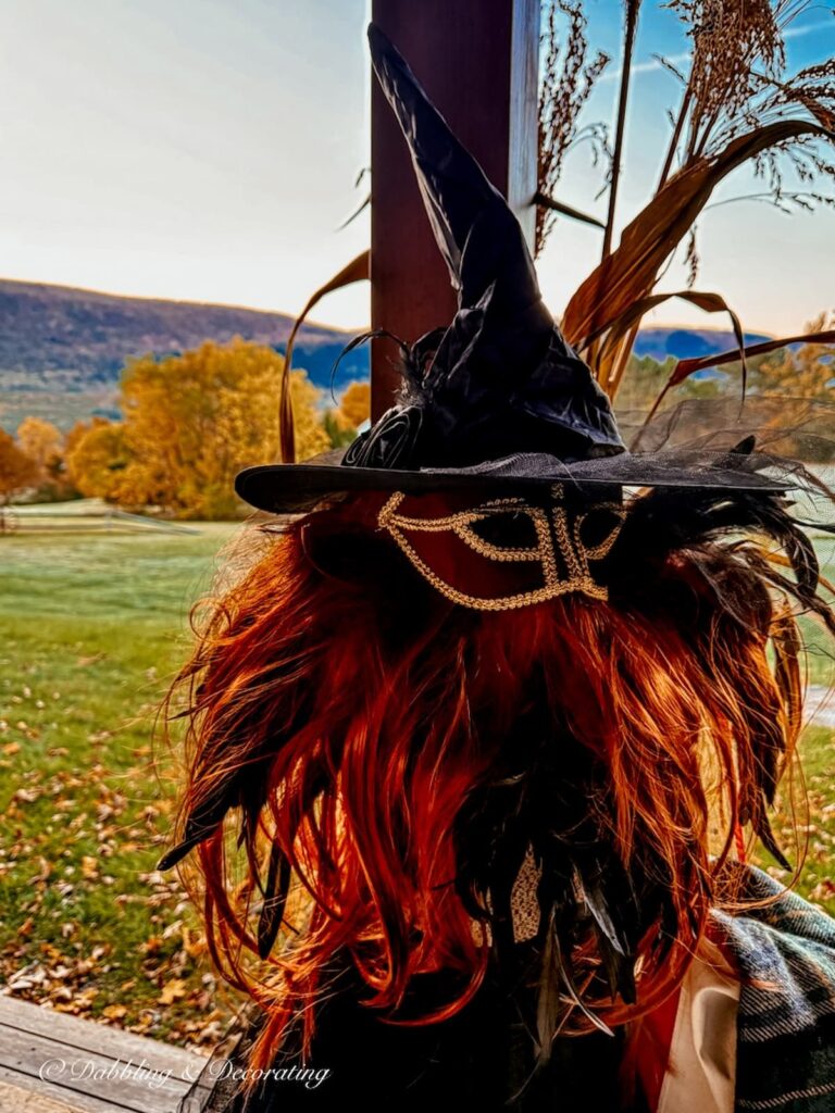 Halloween witch with red wig, mask, black witch's hat and tule skirt in porch decor with fall foliage and mountains in the background.