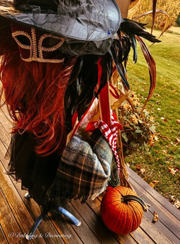 Halloween witch porch ideas with a red wig, mask, black tule skirt, witch's hat pumpkin at feat and an overnight bag with a blanket and red and white stockings on a front porch for Halloween.