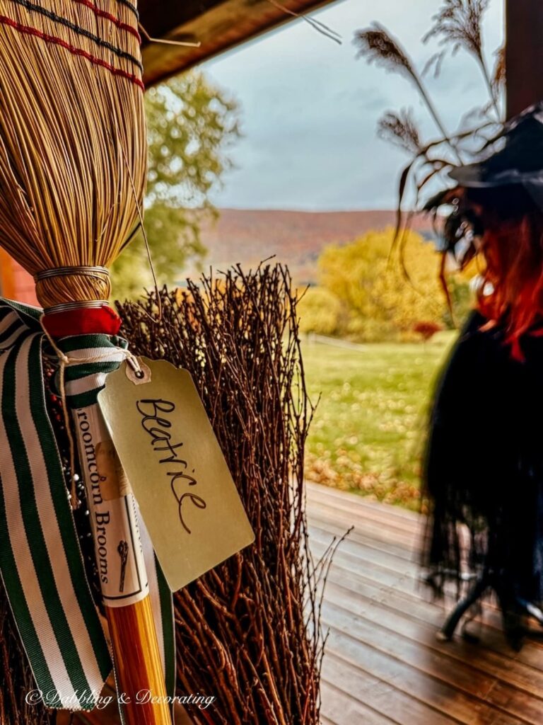 Broom with luggage tag that reads Beatrice on front porch in the mountains during peak foliage with a witch in the background.