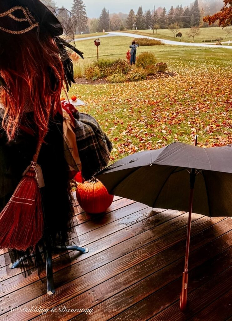 Halloween Witch Porch Ideas with a mannequin dressed witch with broom, open umbrella on porch floor, large pumpkin and peak foliage on a rainy Vermont morning.