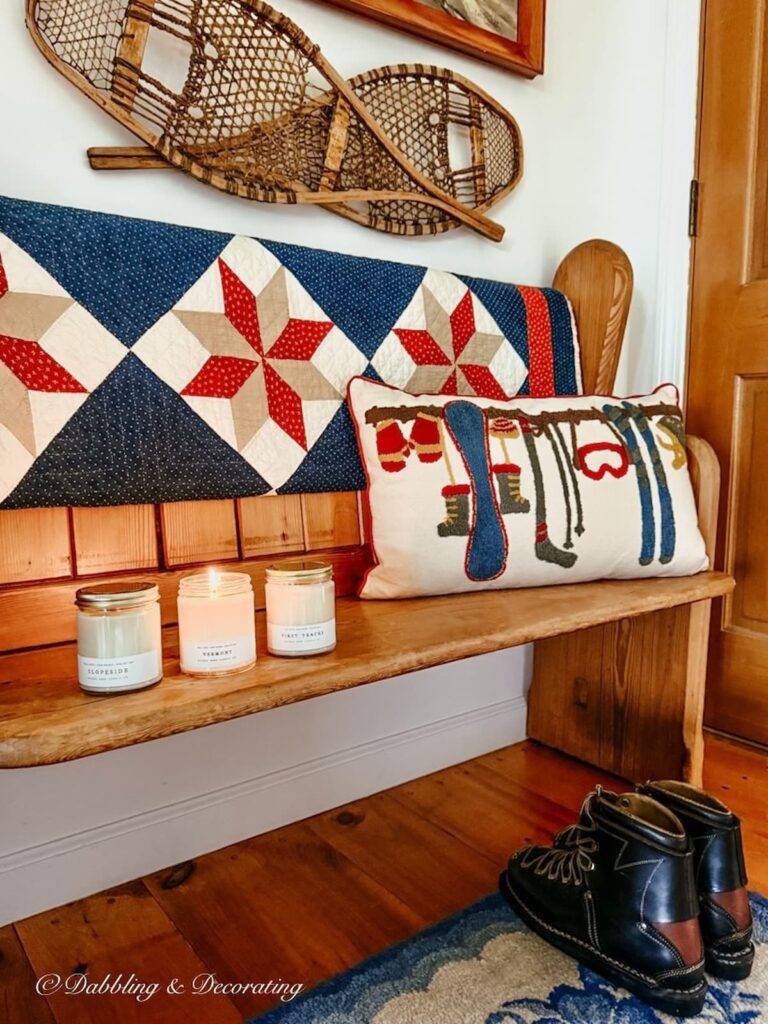 Vintage ski lodge decor in entryway with antique church pew, old quilt, pillow, snowshoes, ski boots and wall painting with Vermont candles by Hayden Rowe Candle Co.