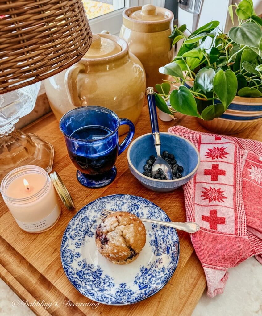 Cozy coffee corner in winter kitchen with vintage plates, mug, alpine towel, lamp on cutting board with yellowware, blueberry muffin on plate and Off Piste Vermont candle.
