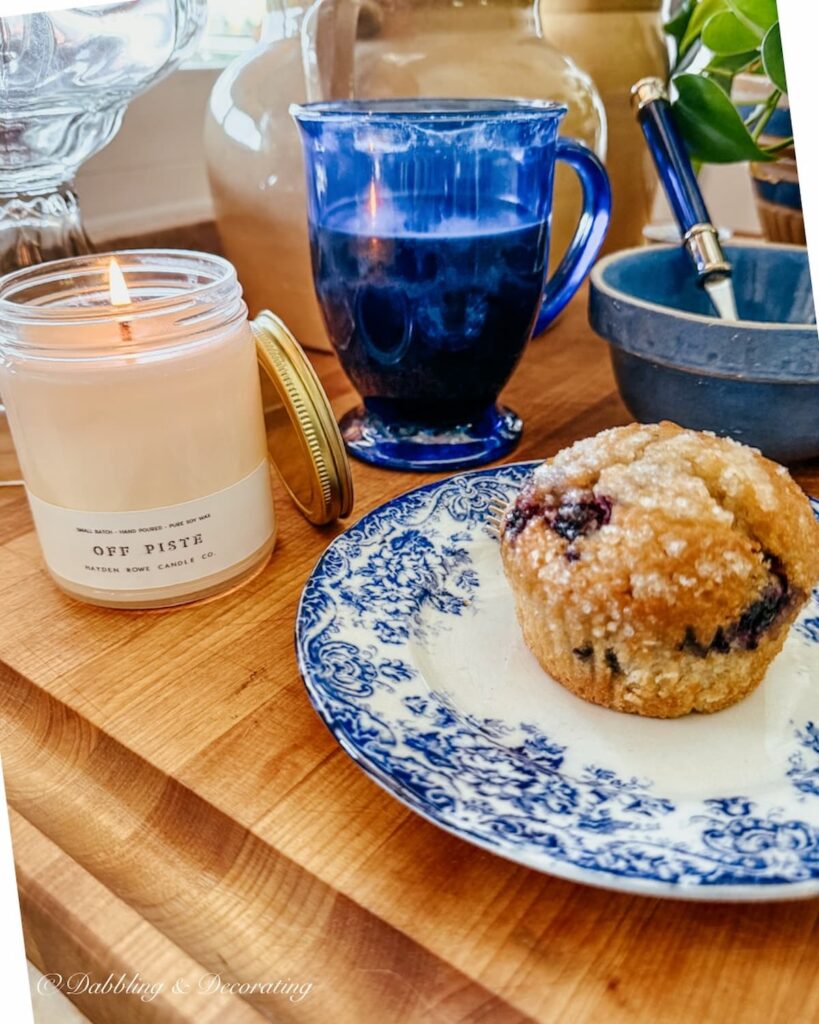 Hayden Rowe Vermont Candle Off Piste on breadboard with morning breakfast with blueberry muffin and blue mug of coffee.