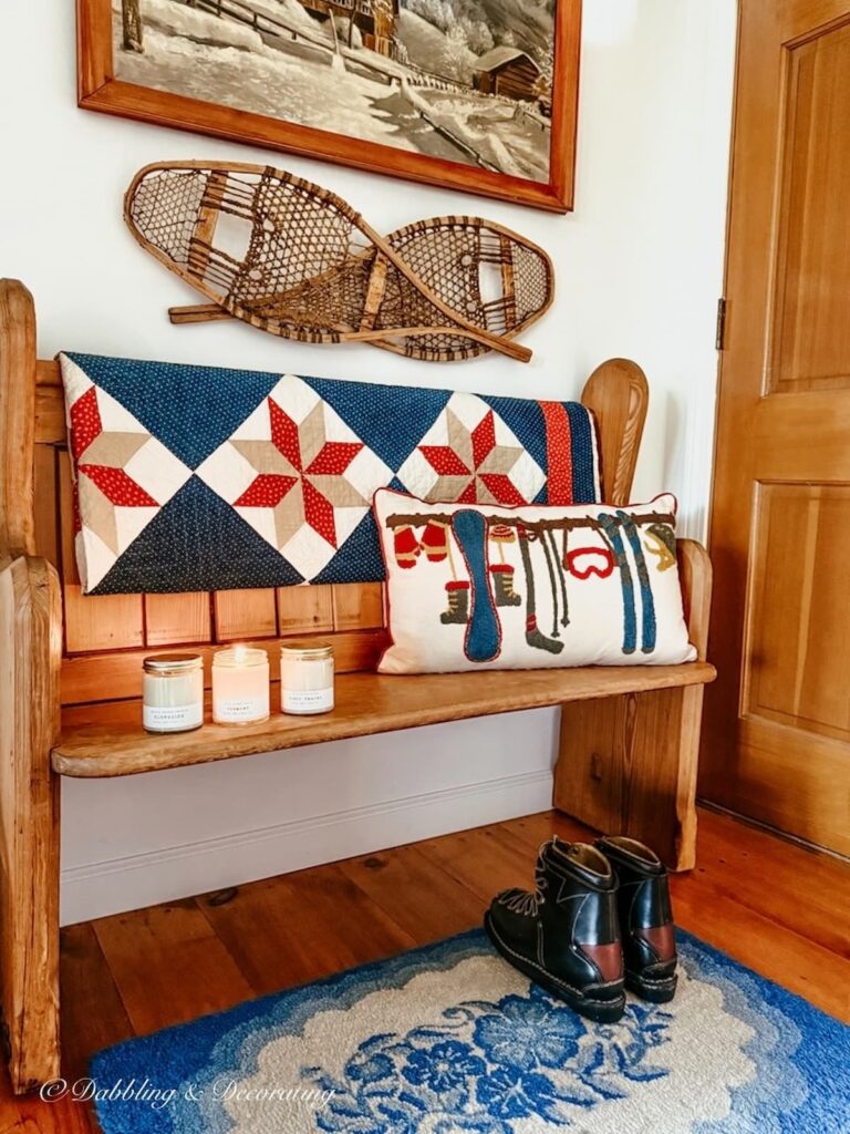 Ski house entryway styled with a vintage church pew, red, white, and blue vintage quilt, ski pillow, old snowshoes and painting on wall, vintage ski boots underneath with blue rug and Hayden Rowe's Vermont Candles to warm the entire space.