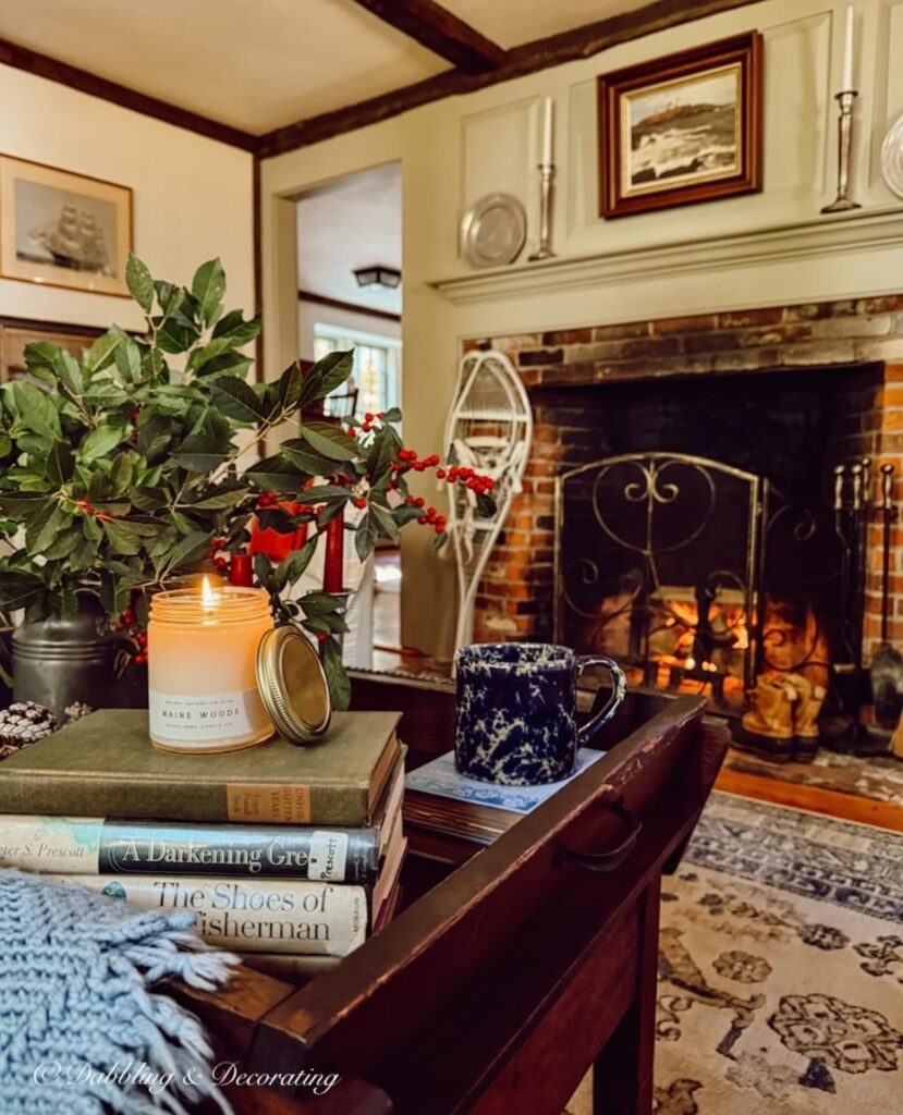 Maine Woods candle burning on a stack of vintage books on a coastal Maine home's antique coffee table with a pitcher of winterberries, coffee mug and fire in the fireplace on a cozy fall day.