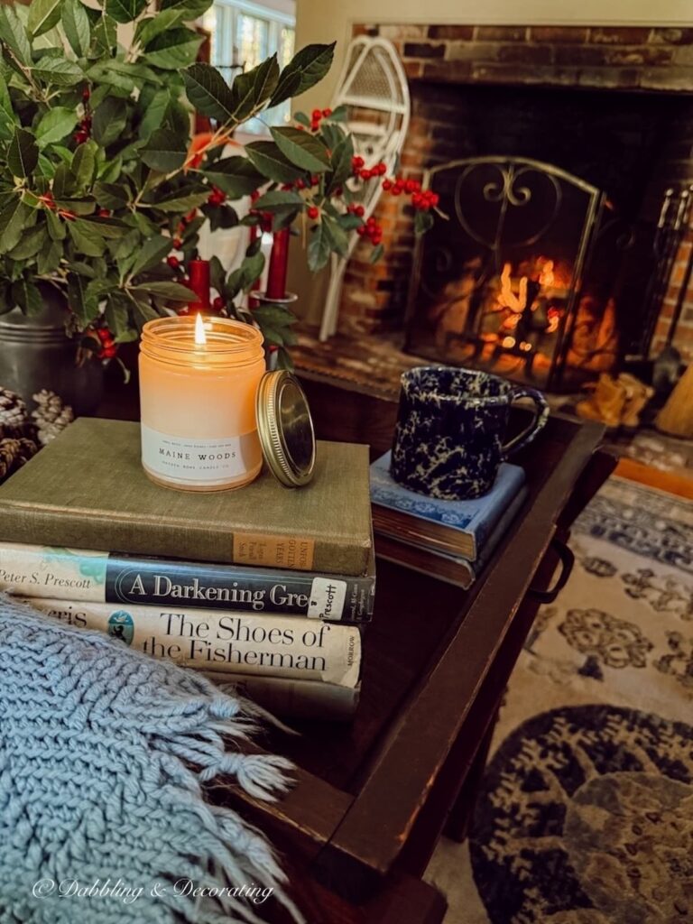 Cozy Maine home's living room with fire in the fireplace and antique coffee table with stacked vintage books, Winterberries in pewter pitcher and Maine Woods candle burning.