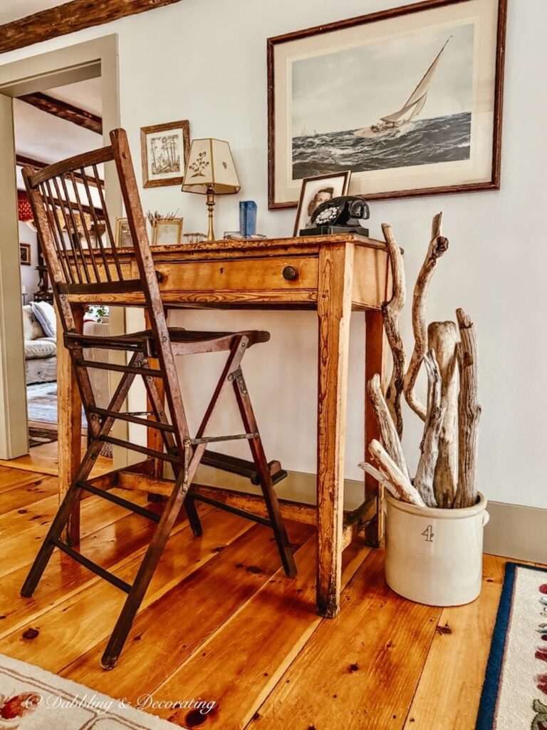 Antique wooden tall desk with vintage umpire chair against a coastal styled wall with a vintage crock on the floor filled with collected driftwood in coastal Maine home.