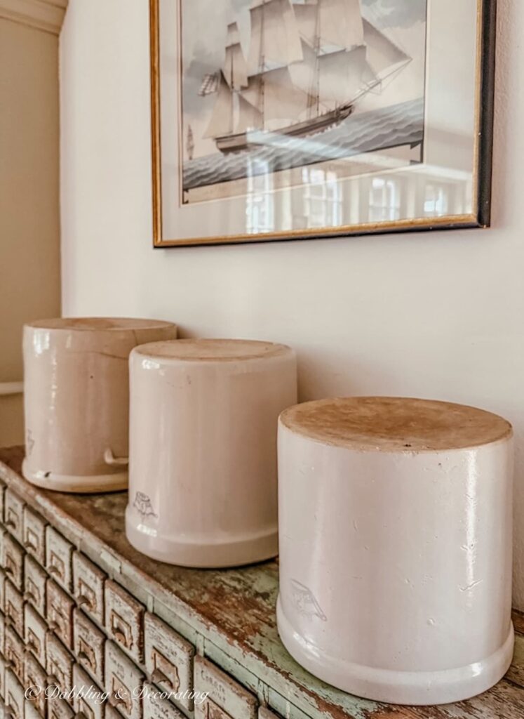 3 Upside down vintage crocks on an antique apothecary cabinet underneath a pastel sailboat wall painting.