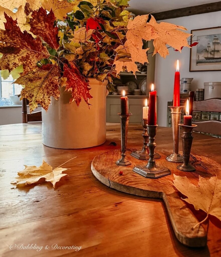 Antique crock filled with foraged fall colorful branches on a round wooden table with a round cutting board with a set of pewter candlesticks with lit candles for a moody table centerpiece vignette in dining room.