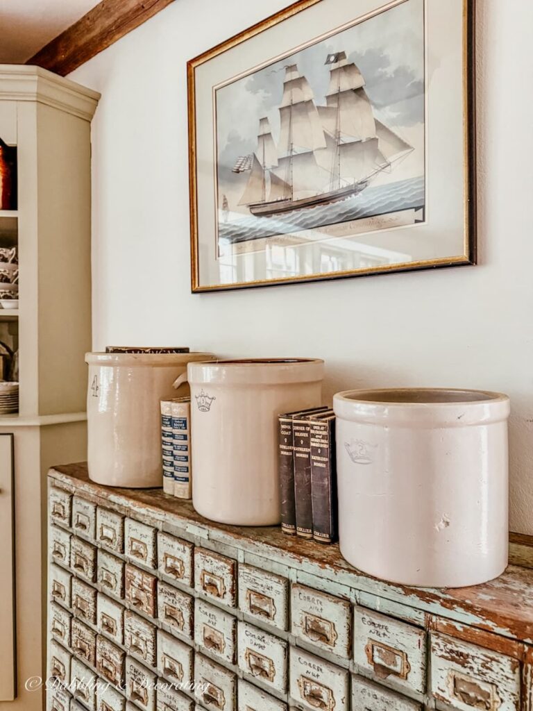 3 vintage crocks on top of an antique apothecary cabinet with vintage books in between the crocks with a pastel sailboat painting hanging on the wall above.