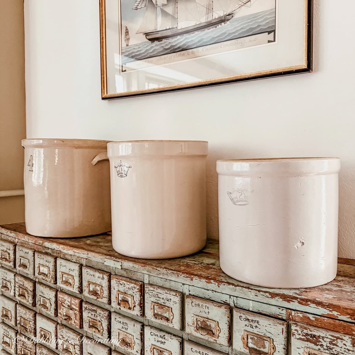 Three vintage crocks displayed on an antique apothecary cabinet with a pastel sailboat painting hanging on the wall above.