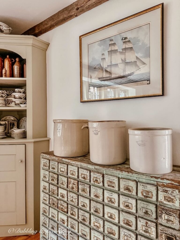 Three Vintage Crocks in different sizes lined up on top of antique apothecary cabinet displayed underneath a pastel sailboat wall painting and beside a corner built-in hutch filled with transferware and old jugs.