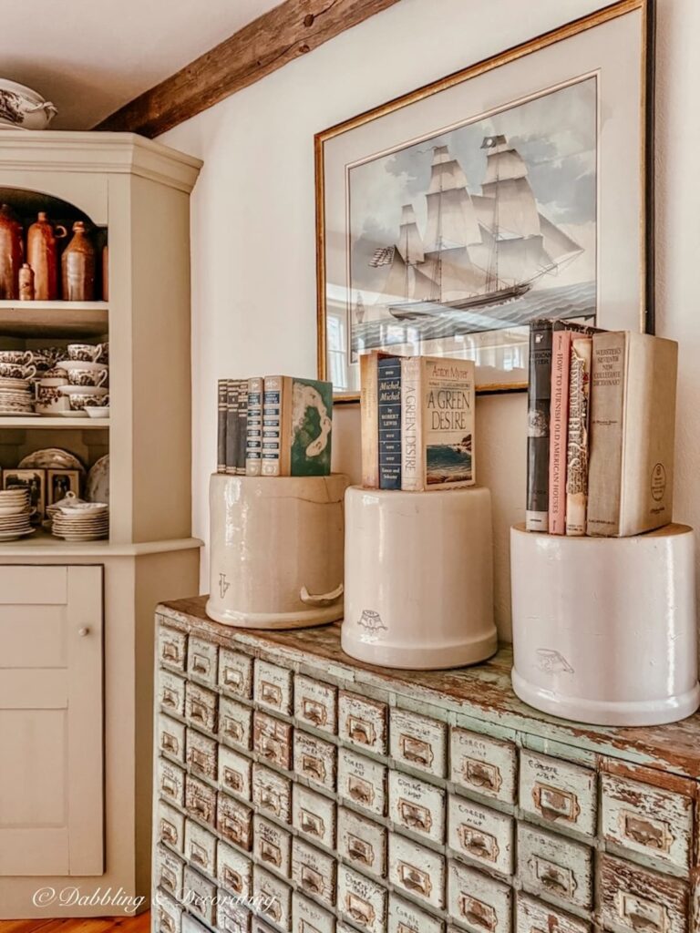 Three vintage crocks in different sizes displayed upside down with vintage books stacked on top of an apothecary cabinet and a pastel sailboat wall painting and corner built-in hutch filled with transferware and old jugs.