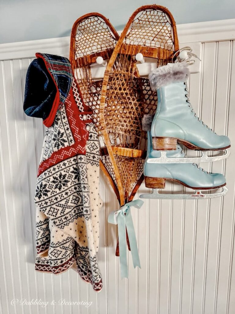 Winter decor vignette with a pair of blue vintage ice skates, pair of vintage snowshoes, thrifted wool ski sweater and old hat on peg board in home entryway on white Wainscoting and blue walls.
